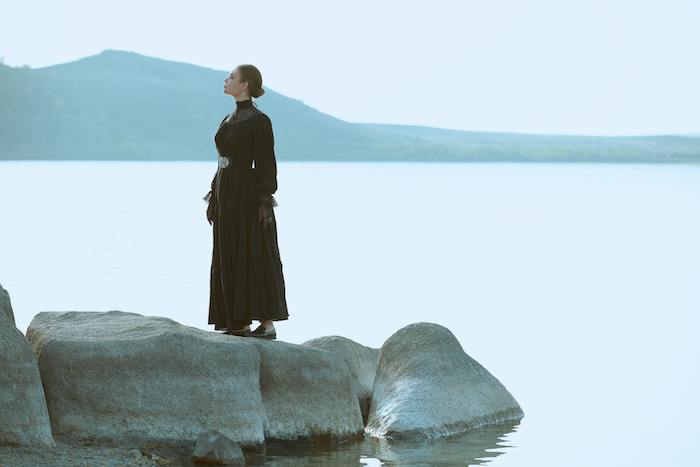 Beautiful, elegant woman in black vintage dress stands by the sea with her back to water pensively and tensely looking into the distance. 19th century novel. Copy space.